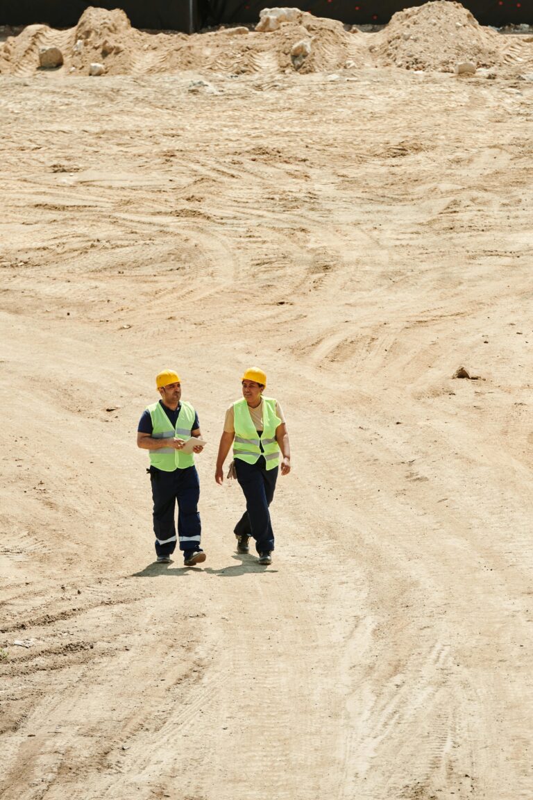 Two workers in safety vests and hard hats walking on a sandy construction site.